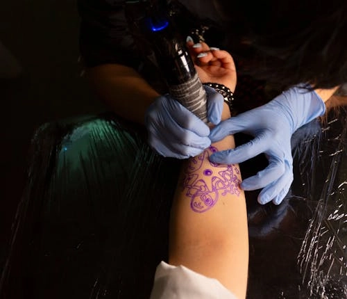 A tattoo artist wearing gloves works on a detailed design on a client’s forearm